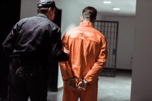 A correctional officer escorts a handcuffed inmate in an orange jumpsuit down a hallway toward a jail cell. The inmate’s back is turned to the camera