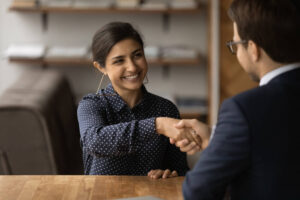 A smiling woman in a polka-dot blouse shakes hands with a man in a suit across a table, suggesting a successful interview or business agreement