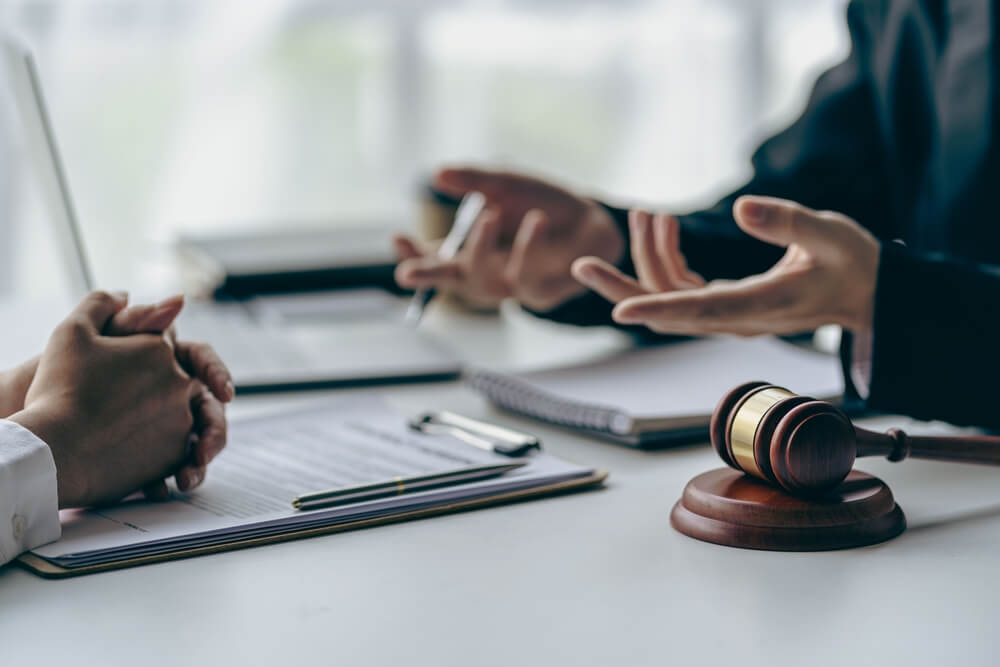 Two people in discussion at a desk with documents, a notebook, and a gavel. One person gestures with open hands, suggesting an explanation or negotiation