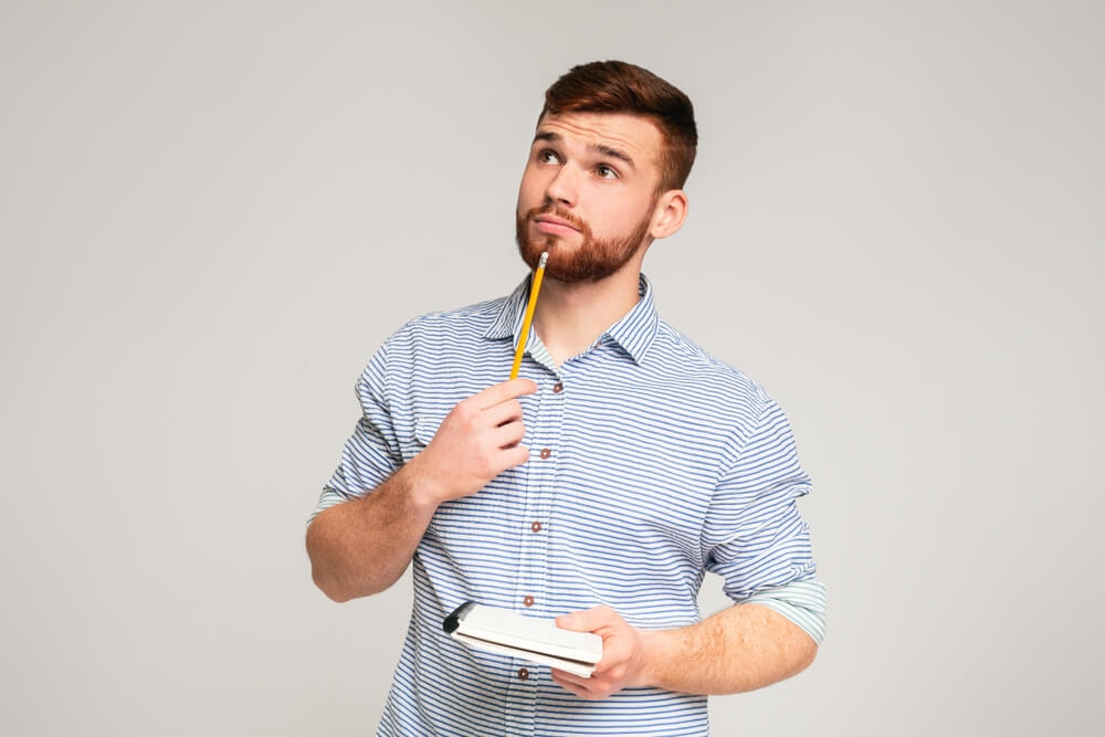 Young man with a thoughtful expression holding a notepad in one hand and a pencil near his chin, looking upward as if contemplating an idea